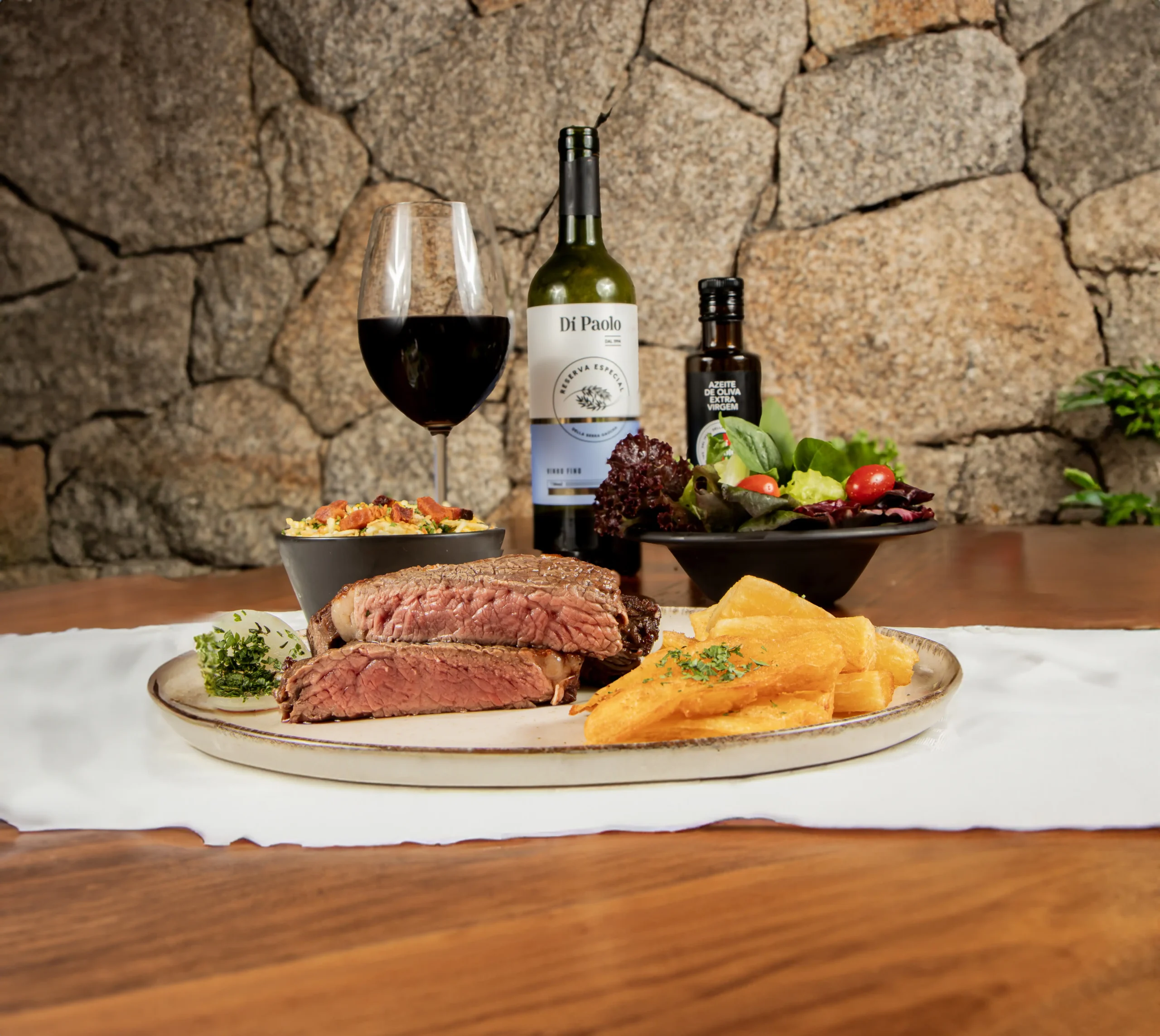 Foto de uma mesa de madeira com um pano branco com um prato grande, duas tigelas pretas, uma taça de vinho, uma garrafa de vinho com a marca do Di Paolo e uma garra de azeite de oliva extra virgem. Dentro do primeiro prato, há dois pedaços de carne, ao ponto, com aparência suculenta e alguns pedaços de mandioca frita ao lado. Nas tigelas, é possível ver uma salada com folhas e tomates cerejas e um acompanhamento para salada. O vinho é tinto e a taça, que é transparente, tem vinho até a sua metade.