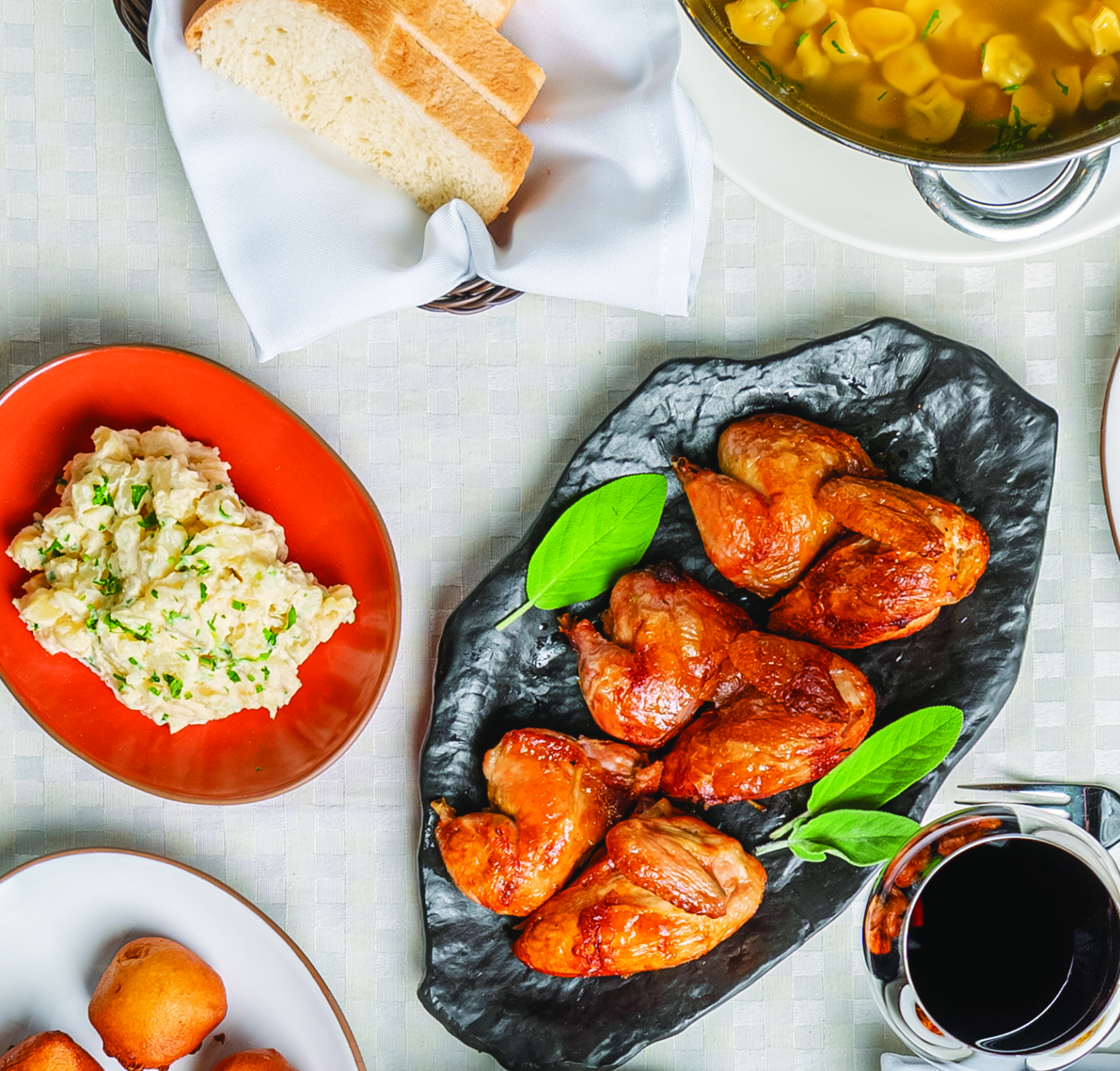 Uma mesa com vários pratos e com uma toalha branca. Pode-se ver com uma visão de cima da mesa: fatias de pães, uma tigela com capeletti, um prato vermelho com maionese de batatas, um prato branco com bordas em tom marrom com bolinhos de queijo, uma bandeja estilizada com 6 pedaços de galetos e uma taça transparente com vinho. 