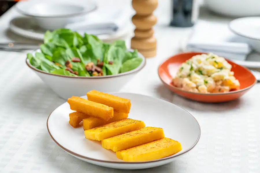 Uma mesa forrada com um pano branco, com destaque para três pratos: um com uma salada de folhas e bacon, outra com arroz e a mais próxima com polenta e queijo fritos.