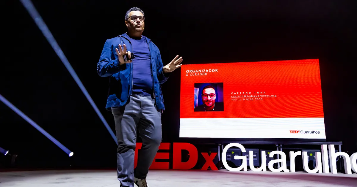 Foto do Caetano Tona, um homem branco, alto, com blazer azul, calça social cinza no palco do TEDx Guarulhos.