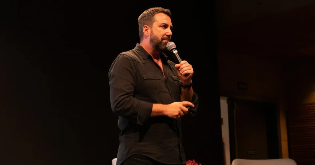 Foto do Carlos Ferreirinha (um homem branco, com cabelo curto e barba bem feita), com camisa preta, segurando um microfone.