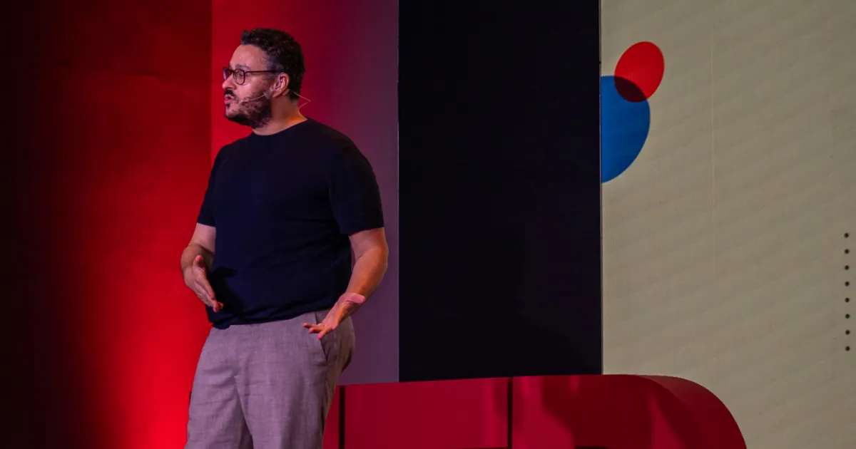 Foto do Caetano Tona, um homem branco e alto, com camiseta preta e calça social cinza, no palco do TEDx Guarulhos