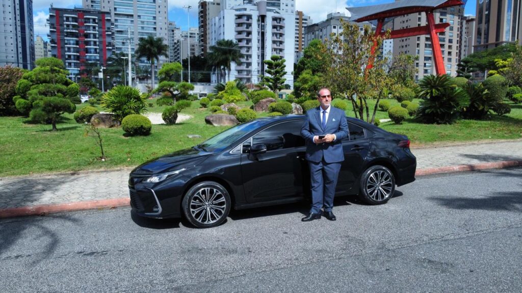 Foto de um motorista executivo encostado em um carro executivo, na cor preta, com vista para o Tori, um monumento japonês que fica em São José dos Campos, São Paulo, Brasil.