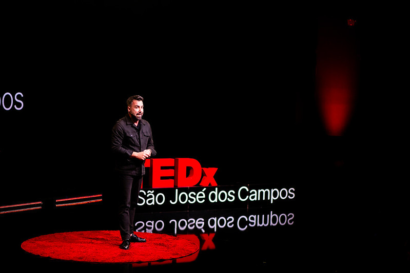 Foto do Carlos Ferreirinha no palco do TEDx São José dos Campos. Um homem branco, vestido de preto, em pé, em cima de um tapete vermelho, em um ambiente escuro e com um letreiro escrito TEDx São José dos Campos ao fundo.