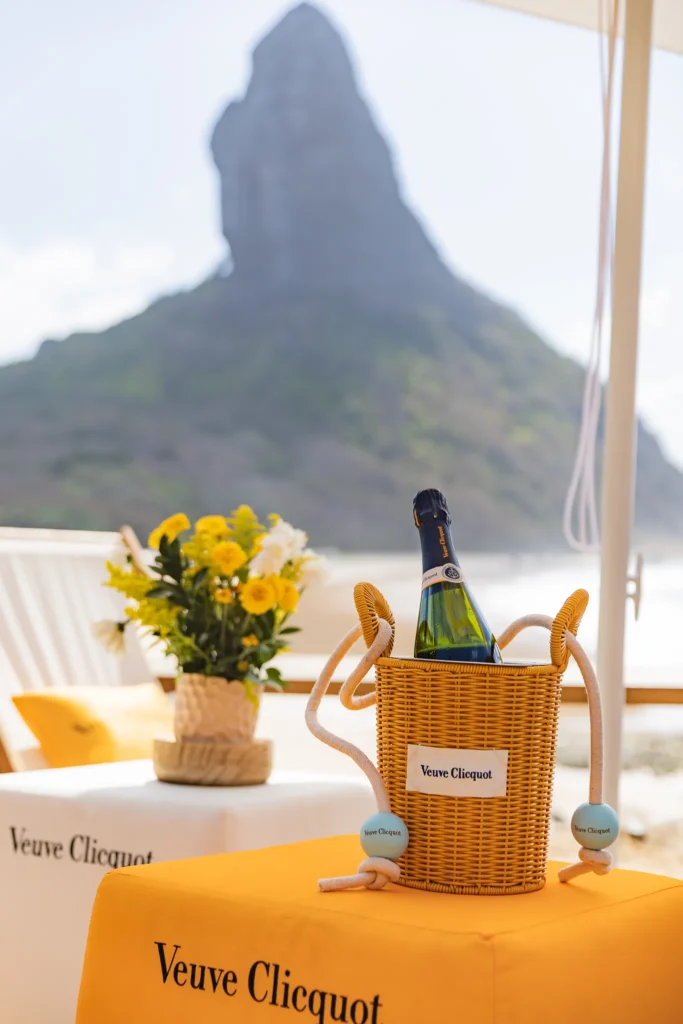 Foto de duas mesas com toalhas escritas "Veuve Clicquot" e um balde com champagne. Ao fundo, uma pedra da praia de Fernando de Noronha