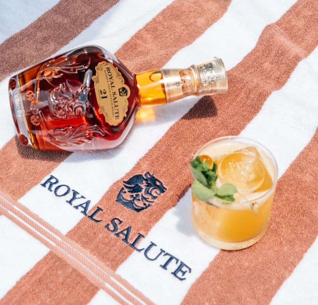 Foto de uma garrafa de Royal Salute 21 anos com um copo do drink Smash Grain ao lado, tudo em cima de uma toalha com o nome do whisky bordado em azul