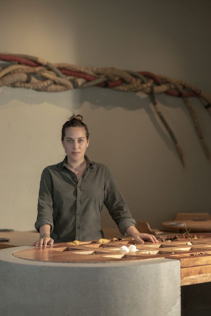 Foto da chef Pía León, uma mulher branca, com cabelos pretos e amarrados, com uma camisa cor cinza escuro, com as mãos em uma mesa de madeira. Nesta mesa, há diversos pratos