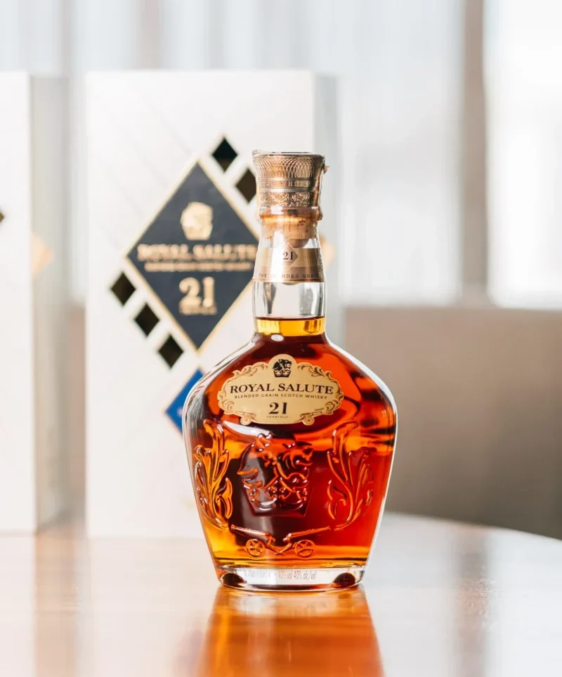 Foto do Royal Salute 21 anos - The Blended Grain, em cima de uma mesa de madeira, com sua caixa branca ao fundo