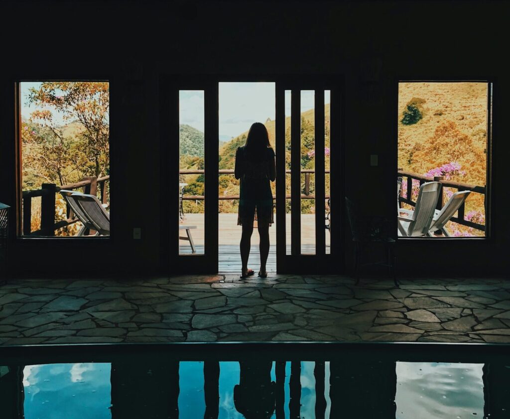 Foto de uma mulher de costas, na porta de uma casa, olhando para as montanhas