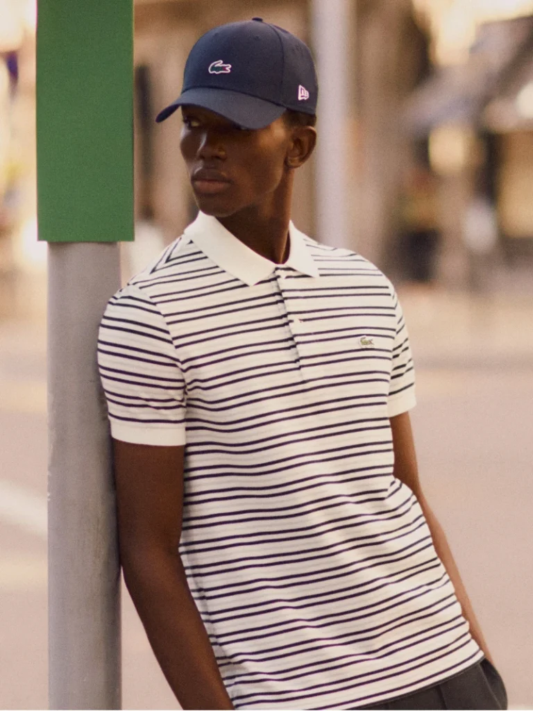 Foto de um homem negro, vestindo uma camiseta listrada e um boné preto da Lacoste e New Era