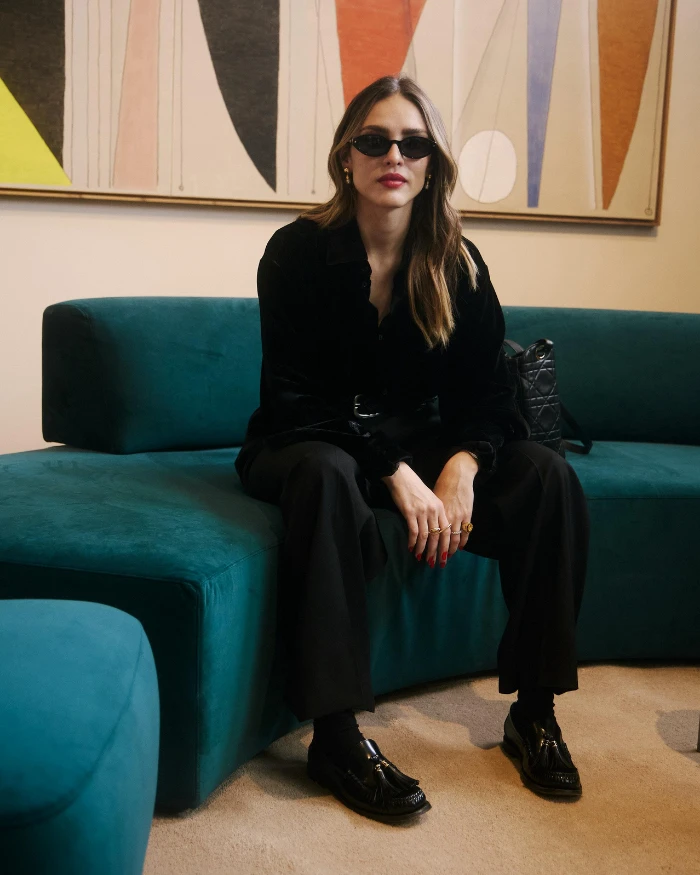 Foto da Isabelle Drummond na SP-Arte 2026, uma mulher branca, com cabelos longos, vestida de preto e sentada em um sofá com cores entre azul e verde