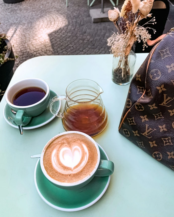 Foto de uma mesa com duas xícara de café, uma com café preto e outra com leite, e parte de uma bolsa da Louis Vuitton ao canto