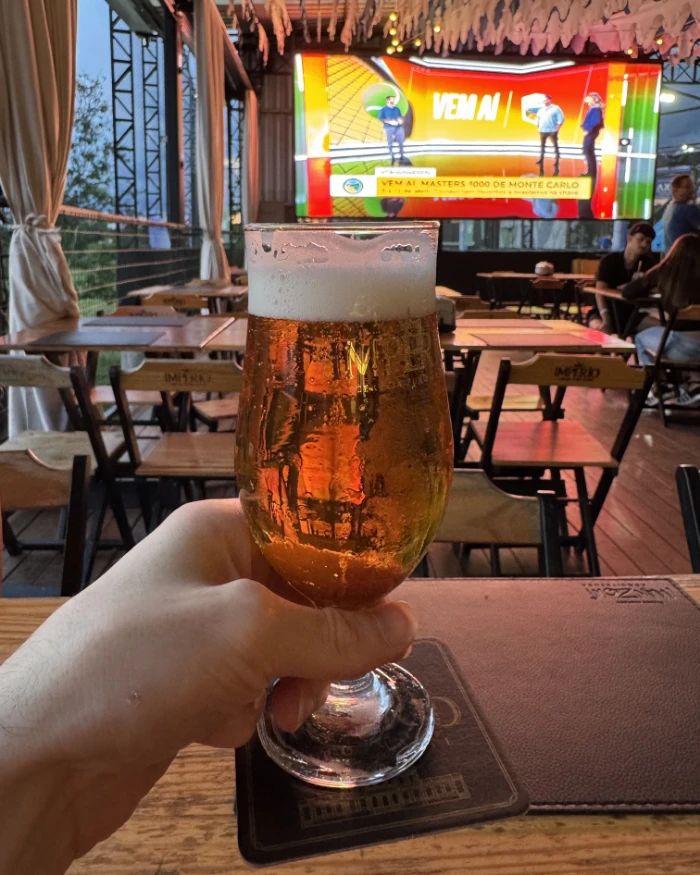 Foto de uma pessoa segurando um copo de Chopp de frente para um telão de led no Arpoador Posto012 em São José dos Campos