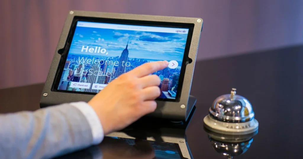 Foto de uma pessoa tocando na tela de um sistema de check-in em um tablet. Ao lado do tablet, há uma campainha de apertar