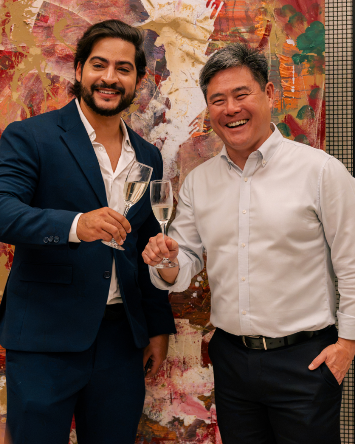 Foto de Lucas Toledo, homem branco, cabelos pretos, camisa branca e blazer azul, ao lado de Marcelo Midorikawa, homem branco, traços asiáticos, camisa branca e calça preta. Ambos estão sorrindo e segurando uma taça de espumante nas mãos