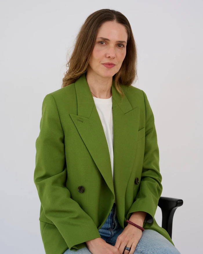 Foto de Giovanna Meneguel, uma mulher branca de cabelos compridos e castanhos, com um blazer verde, camiseta branca e calça jeans clara