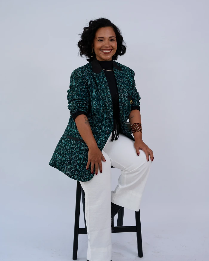 Foto de Noanny Maia, uma mulher negra, com blazer em tons verdes e calça branca