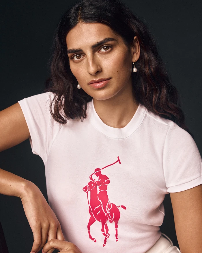 Foto de Saffron Vadher, uma mulher de cabelos pretos, vestindo uma camiseta rosa clara com o símbolo rosa da Ralph Lauren estampado no meio