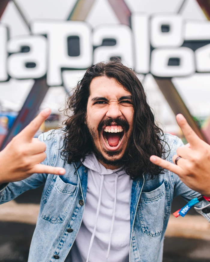 Foto de um homem branco de cabelos longos fazendo o símbolo do rock and roll nas mãos com o letreiro do Lollapalooza no fundo
